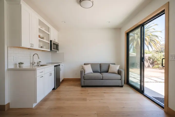 The interior of a small modern ADU living area with a compact kitchenette along one wall in white cabinets with a quartz counter and a small sink