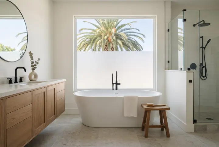 A primary bathroom with a white freestanding soaking tub placed in front of a large frosted-lower picture window with a palm tree visible above the frosting