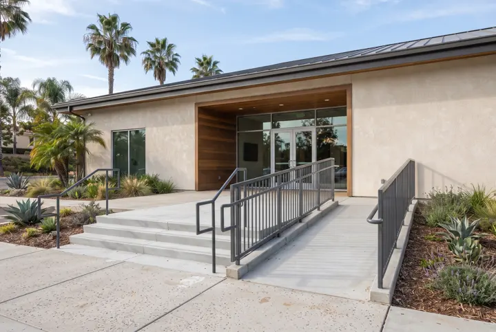 New light-gray concrete entry steps and a gently sloped concrete ramp with a painted steel railing at the entrance of a single-story community building with stucco walls