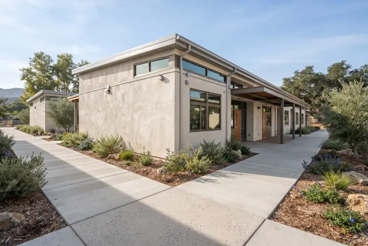 A concrete flatwork walkway around the perimeter of a single-story stucco school building with clean control joints and planter beds with drought-tolerant plants
