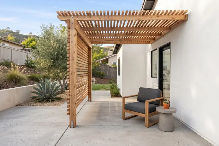 A modern slatted cedar pergola with horizontal wood slats casting striped sunlight over a clean gray concrete patio