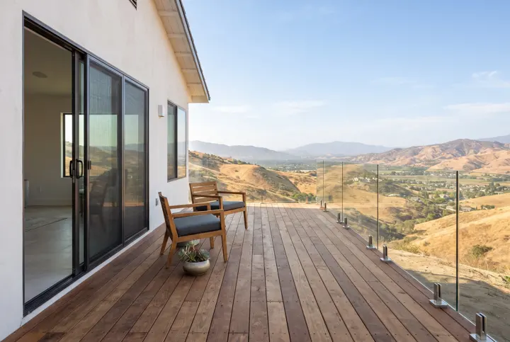 A elevated wood deck with a frameless glass railing overlooking dry golden California hills and a sunny valley