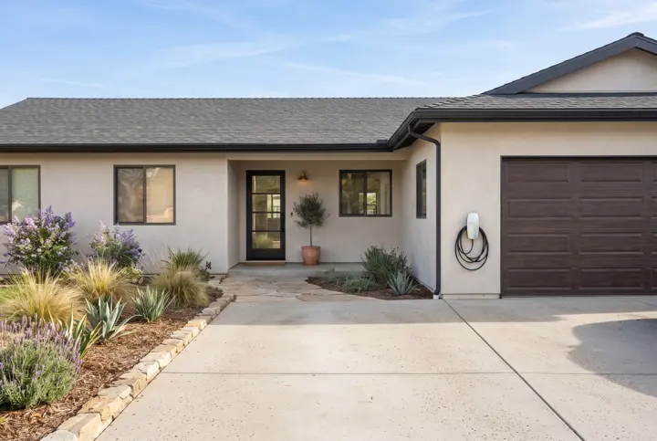A modern stucco home with a clean broom-finish concrete driveway and a small white wall-mounted electric vehicle charger with a neatly coiled cable on the wall beside the garage door