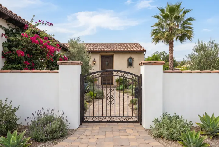 A smooth white stucco wall with a cap and a black wrought-iron gate set between two stucco pillars