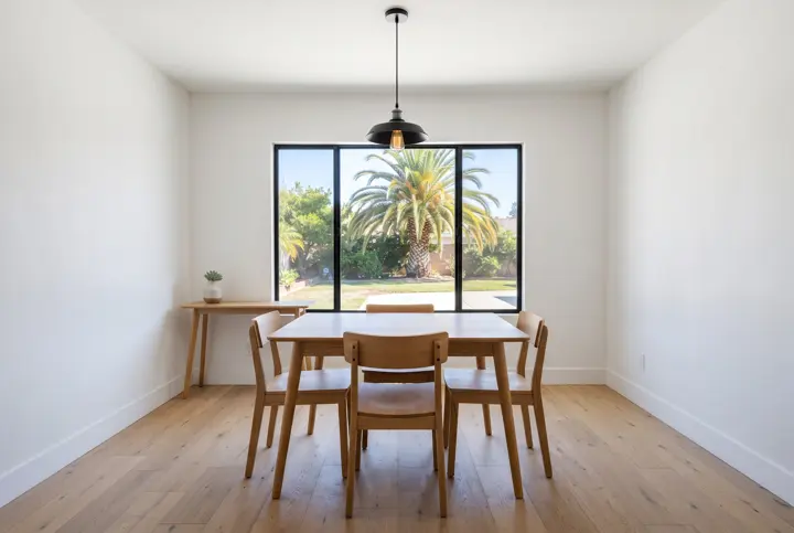 A dining room with a large picture window with black frames looking out to a sunny yard and palm tree