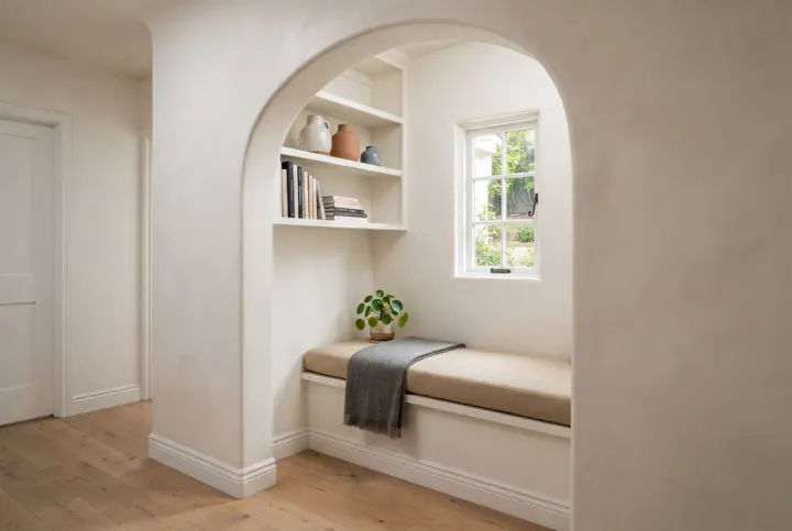 A reading nook built into a bright hallway alcove with a plaster arch, an upholstered bench seat