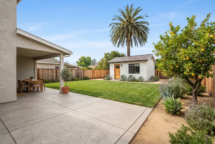 A wide backyard view with a green lawn and a stamped-concrete patio in the foreground and a small modern white guest house with a wood door in the far distance under a palm tree