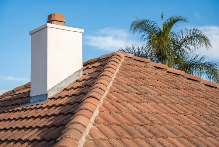 A close-up detail of a new terracotta tile roof ridge with clean mortared ridge caps and neat metal flashing where the roof meets a white stucco chimney