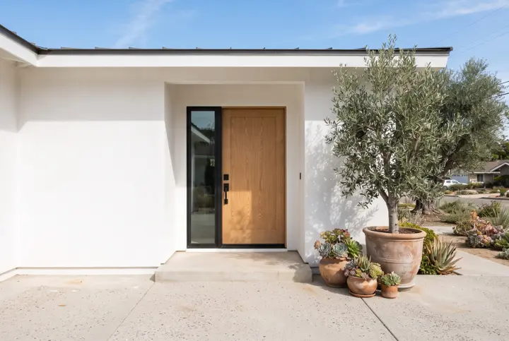 A modern solid wood front door with a black handle set in a clean white stucco facade with a smooth blank wall